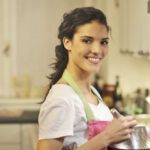 Culinary Delights - Side view of cheerful female in apron and casual t shirt standing in modern kitchen and mixing ingredients with whisk in stainless bowl while preparing dough