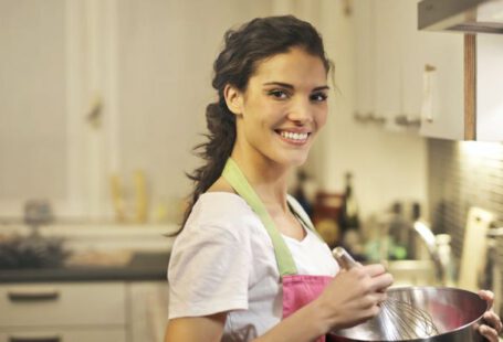 Culinary Delights - Side view of cheerful female in apron and casual t shirt standing in modern kitchen and mixing ingredients with whisk in stainless bowl while preparing dough